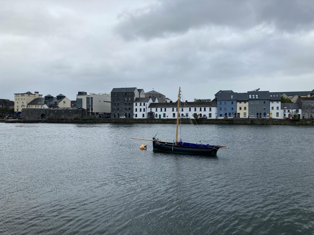 A lone sailboat on tranquil water with a backdrop of urban buildings under a cloudy sky.